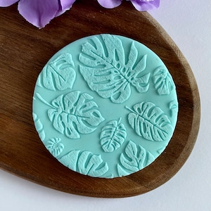 May include: A light blue silicone mold with a raised design of tropical leaves. The mold is circular and has a smooth surface.
