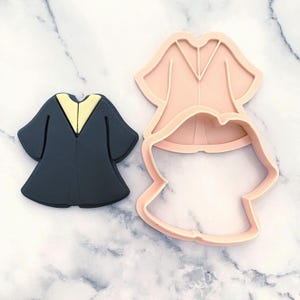 Puede incluir: Un cortador de galletas de toga de graduación azul marino oscuro con un cuello amarillo está junto a un cortador de plástico rosa en forma de toga de graduación. El cortador de galletas es una herramienta para hornear.