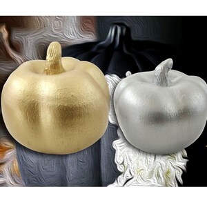 May include: Two decorative pumpkins, one gold and one silver, are displayed against a dark background. The pumpkins are smooth and have a glossy finish.