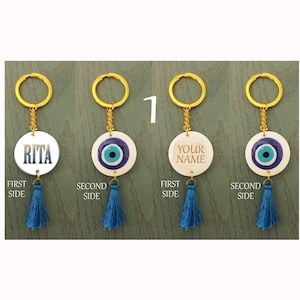 Op de afbeelding: Vier sleutelhangers met een blauwe boze oog-hanger aan de ene kant en een gepersonaliseerde optie aan de andere kant. De gepersonaliseerde opties omvatten een blauwe boze oog, de naam "RITA" en de tekst "YOUR NAME".