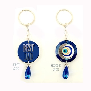 Puede incluir: Un llavero azul y blanco con una cadena plateada y un dije que dice "Best Dad" en un lado y un dije azul de ojo turco en el otro.