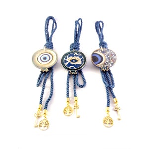 Può includere: Tre ciondoli portafortuna blu e oro con nappe e chiavi. Ogni ciondolo presenta un design diverso: un occhio blu, un occhio blu su sfondo bianco e un occhio blu su sfondo bianco con una mezzaluna.