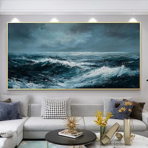 Könnte beinhalten: Großformatiges Ölgemälde, das eine stürmische Meereslandschaft zeigt. Das Kunstwerk zeigt dunkelblaue und weiße Wellen unter einem bewölkten Himmel, mit einem goldenen Rahmen. Das Gemälde hängt über einem grauen Ecksofa in einem Wohnzimmer.