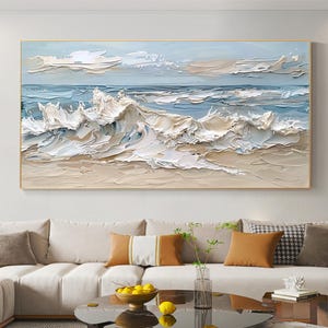 Op de afbeelding: Een ingelijst olieverfschilderij dat een zeegezicht weergeeft met getextureerde golven in wit, beige en blauw. Het kunstwerk heeft een gouden lijst en wordt tentoongesteld boven een beige bank met kussens. Het schilderij roept de oceaan op.