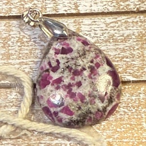 May include: A teardrop-shaped pendant featuring a mottled pattern of deep magenta and white stone fragments. The pendant is topped with a polished silver-toned metal bail, designed to hold a chain or cord for wear as a necklace accessory.