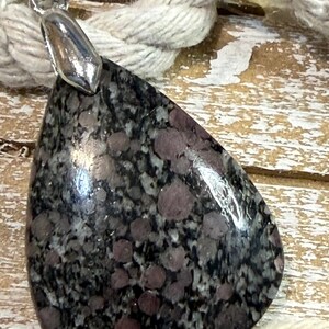 May include: A teardrop-shaped pendant with a polished, speckled stone in shades of gray and maroon. The pendant has a silver-colored bail. A thick, off-white rope is in the background.