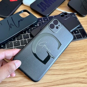 May include: A collection of black phone cases with personalised names, including "Chloe", "James", "Grace", and a heart. The cases are made of a smooth, matte material and have a sleek, modern design.