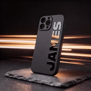 May include: A matte black smartphone case featuring the name JAMES in large, vertical, reflective silver capital letters. The case includes a square camera cutout with three circular lens openings and side button cutouts on the right edge.