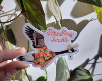 May include: A sticker of a cartoon goose wearing a floral shirt and a straw hat, flying with the text "Heading South" in red.