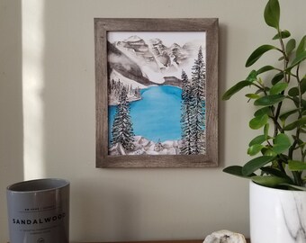 May include: A framed watercolor painting of a mountain lake scene with snow-capped mountains in the background and two pine trees in the foreground. The lake is a vibrant blue color.