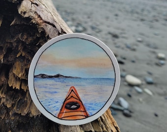 May include: A round sticker featuring a watercolour illustration of a kayak paddling towards a distant shoreline. The sky is a soft blue with a hint of orange and the water is a deep blue with whitecaps.