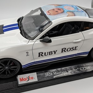 May include: White 2020 Ford Mustang Shelby GT500 diecast model car with blue racing stripes and a personalized photo of a baby on the roof. The car has the name "Ruby Rose" printed on the side. The car is on a black display base with the Maisto logo.