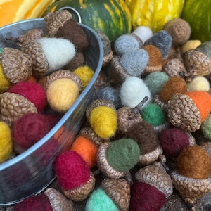 May include: A collection of felt acorns in various colors, including red, yellow, green, brown, and white, are displayed in a metal bucket and scattered on a wooden surface.