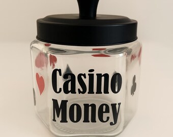 May include: A clear glass jar with a black lid and handle, labeled "Casino Money" in bold black letters. Red hearts and spades decorate the sides. A decorative container for game night.