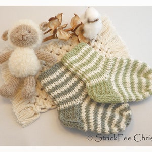 Könnte beinhalten: Ein gestricktes Lamm-Spielzeug und zwei Paar Babysocken. Die Socken sind grün und weiß gestreift. Das Lamm ist beige und weiß. Die Artikel liegen auf einer cremefarbenen Makramee-Spitze mit Baumwollstielen.