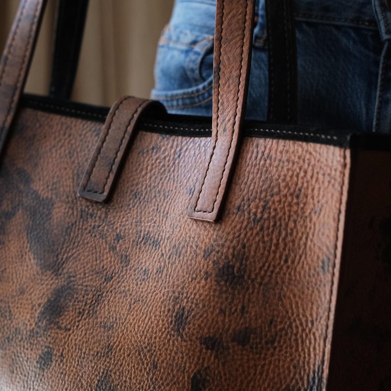 May include: A brown leather tote bag with a textured surface and black accents. The bag has two handles and a rectangular shape. The leather has a mottled pattern with darker brown spots.