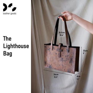 May include: A brown leather tote bag, labeled "The Lighthouse Bag," with black handles and a buckle closure. The bag measures 13.3 inches wide, 9 inches high, and 3.1 inches deep. Handle drop is 10.4 inches.
