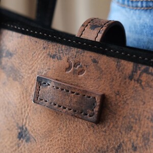May include: Close-up of a brown leather tote bag with a black suede trim. The bag features a textured surface with dark spots, a stitched leather handle, and a rectangular leather tag with stitched details. A small logo is embossed on the leather.