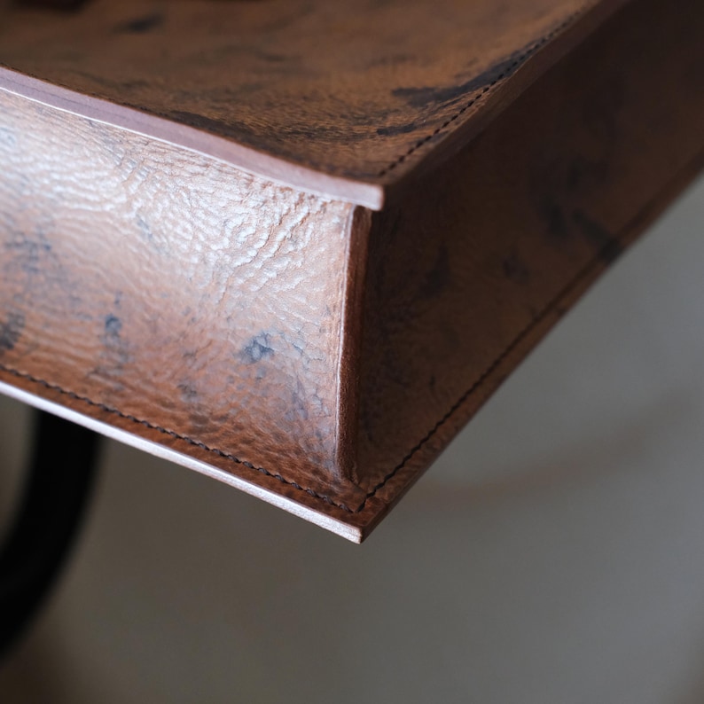 May include: Close-up of a handcrafted, rectangular leather tray. The tray is a rich brown color with a textured surface and visible stitching along the edges. The leather appears to have a natural, aged look.