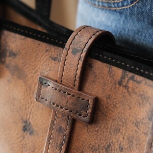 May include: Close-up of a brown leather tote bag with a handle and a rectangular strap. The leather has a distressed appearance with dark spots. The handle is stitched with a lighter brown thread.