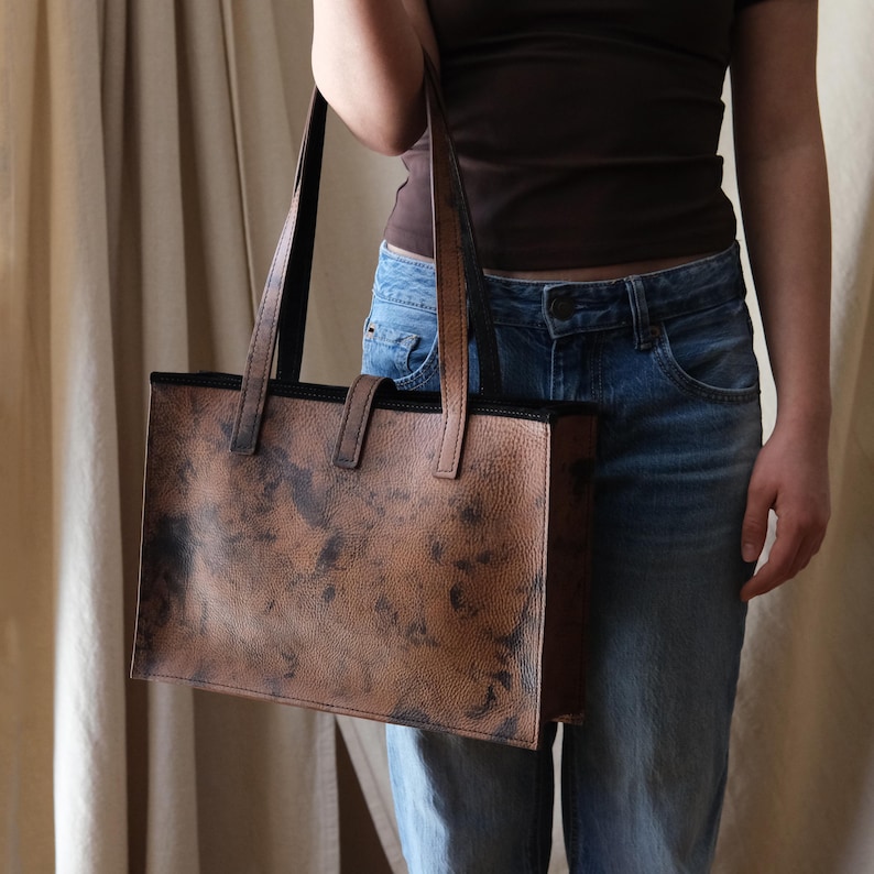 May include: A brown leather tote bag with a textured finish and black trim. The bag has two long handles and a small handle on the front. The bag is rectangular and appears to be empty.