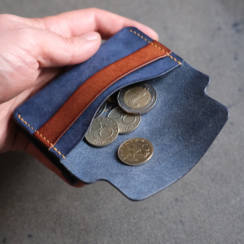 May include: A blue leather wallet with a brown leather flap and a stitched edge. The wallet is open and has several gold coins inside.
