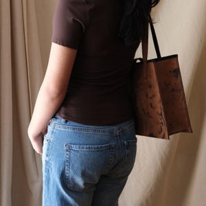 May include: A brown leather tote bag with a textured, mottled appearance. The bag has a rectangular shape with a black trim and handles. The bag is carried over the shoulder.