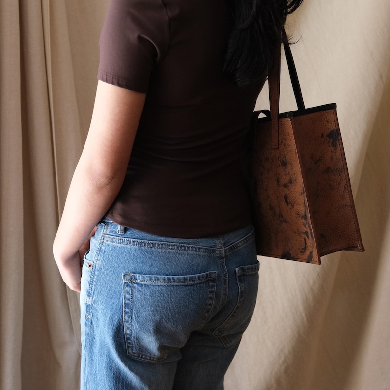 May include: A brown leather tote bag with a textured, mottled appearance. The bag has a rectangular shape with a black trim and handles. The bag is carried over the shoulder.