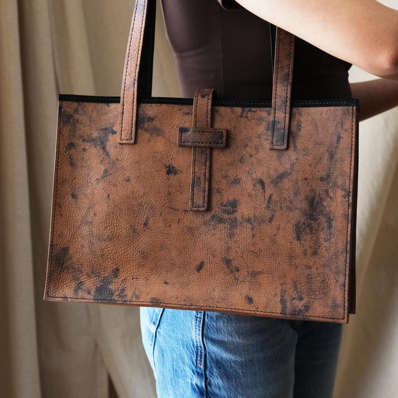 May include: A brown leather tote bag with a rectangular shape and two handles. The bag features a unique, distressed pattern with dark spots and a strap closure. The bag is held by a person wearing blue jeans.