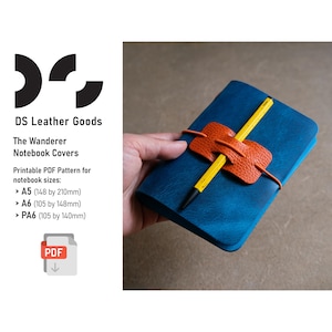 Pode incluir: Um caderno de couro azul com um porta-canetas de couro laranja e um lápis amarelo. O caderno é segurado por uma mão. O texto na imagem diz "DS Leather Goods" e "The Wanderer Notebook Covers".