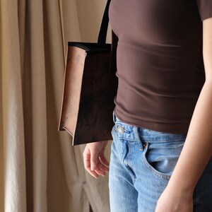 May include: A brown tote bag with a textured leather-like appearance and black straps. The bag is carried over the shoulder. The person is wearing a brown top and blue jeans.