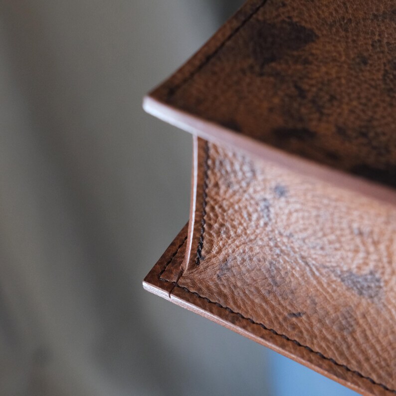 May include: Close-up of a brown leather item with a textured surface and visible stitching. The item appears to be a corner of a larger piece, possibly a bag or wallet, with a detailed pattern.