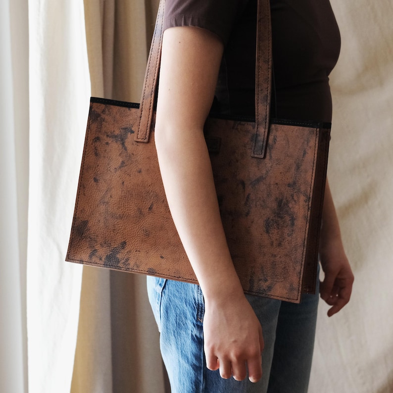 May include: A brown leather tote bag with a rectangular shape and two handles. The bag has a distressed, mottled appearance with dark patches. The bag is being held by a person wearing a brown shirt and blue jeans.
