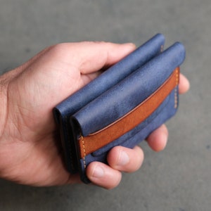 May include: A blue leather wallet with a brown leather accent and stitching. The wallet is folded in half and held in a hand.
