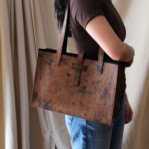 May include: A brown leather tote bag with a rectangular shape and a distressed finish. The bag has two handles and a strap closure. The interior is black. The bag is being carried by a person.