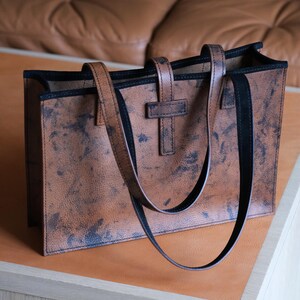 May include: A brown leather tote bag with black trim and handles. The bag has a textured, distressed appearance with dark blue-black accents. The handles are attached with a T-shaped leather piece. The bag is rectangular and sits on a light brown surface.
