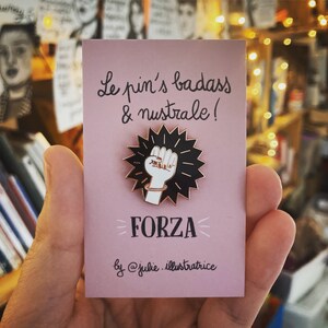 Peut inclure: Une carte rose avec le texte "Le pin's badass & nuistrale !" et "FORZA" en lettres noires. Une épingle en émail noir et blanc avec un poing levé est sur la carte. Le texte "by @julie.illustratrice" est en bas de la carte.