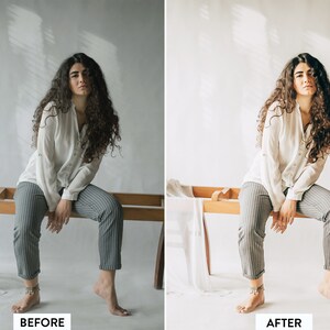 May include: A woman with long, curly brown hair sits on a wooden bench. She is wearing a white button-down shirt and striped trousers. The background is a white wall with shadows.