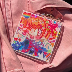 Może przedstawiać: Kolorowy, błyszczący brelok z postacią z kreskówki o pomarańczowych włosach, czerwonej kokardce i tęczowym sercem. Brelok jest otoczony gwiazdami, sercami i nutami.