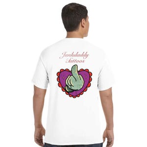 Könnte beinhalten: Ein weißes kurzärmeliges T-Shirt mit einer Grafik auf der Rückseite. Das Design zeigt den Text Junkdaddy Tattoos in roter Schreibschrift über einem violetten Herz mit rotem Wellenrand, das eine grüne Hand mit einer Geste enthält.