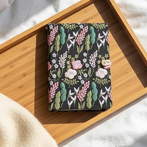 Puede incluir: Un diario negro con un estampado floral en rosa, verde y blanco, apoyado en una bandeja de madera marrón claro. El diario tiene un cierre de botón. La bandeja está sobre una sábana blanca con una manta beige.