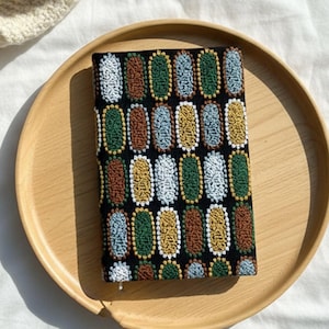 Pode incluir: Uma capa de caderno preta com um padr&atilde;o bordado colorido de ovais verticais em branco, verde, amarelo, castanho e azul. O caderno est&aacute; numa bandeja redonda de madeira.
