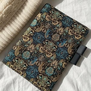 Op de afbeelding: Een zwarte, bedrukte boekomslag met een bloemmotief in teal, goud en blauw. De omslag heeft een zwarte leren bandsluiting. De omslag ligt op een wit oppervlak naast een crèmekleurige kabelgebreide trui.