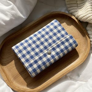 Peut inclure: Un portefeuille à motif vichy bleu et blanc avec une fermeture à bouton-pression argentée repose sur un plateau ovale en bois. Le plateau est brun clair et posé sur une surface blanche, avec une couverture en tricot crème en arrière-plan.