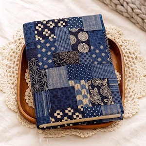 Puede incluir: Una cubierta de libro de patchwork azul con varios patrones geométricos y florales. La cubierta está hecha de diferentes tonos de tela azul y descansa sobre una bandeja de madera con un tapete de encaje blanco debajo. Una cinta blanca de marcador es visible.