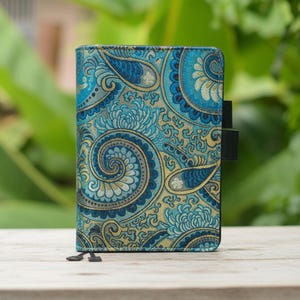 Puede incluir: Un diario con un estampado de cachemira azul y dorado y un cierre elástico negro. El diario está sobre una superficie de madera con un fondo verde borroso. La portada presenta un diseño floral detallado.