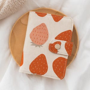 May include: A cream-colored wallet with a strawberry pattern. The wallet features a repeating design of large, orange strawberries with white seeds. The wallet is closed with a button clasp. The wallet is on a wooden plate.