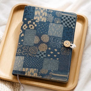 Puede incluir: Una cubierta de diario de tela azul con un diseño de patchwork de patrones florales y geométricos. La cubierta se asegura con un botón de madera y un lazo de cordón blanco. Un dije decorativo de metal cuelga de un marcapáginas de cinta. El diario descansa sobre una bandeja de madera.