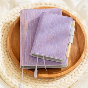 May include: Two lavender journals with a wood-grain effect are stacked on a wooden tray. A pen is attached to the smaller journal. The tray rests on a cream-coloured, knitted placemat, creating a cosy, organised aesthetic.