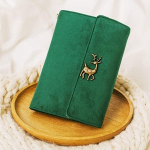 Puede incluir: Una cartera verde con un emblema de ciervo dorado. La cartera está hecha de un material suave similar al ante y tiene un cierre de solapa. La cartera está sobre una bandeja de madera y una manta de punto blanca.